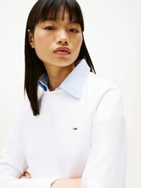 Sudadera blanca con cuello redondo y mangas largas, que presenta un pequeño logo. Llevada sobre una camisa de cuello azul claro, con textura de tela suave.