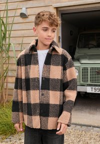 Chaqueta de forro polar a cuadros en tonos marrones y negros, con grandes cuadrados, un cuello y botones en el frente. Lleva puesta sobre una camiseta blanca con pantalones oscuros.