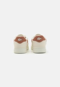 Sneakers Lacoste con una toma beige caratterizzata da un design testurizzato e accenti di colore ruggine sul tallone. Lacci bianchi e dettaglio del logo sul tallone.