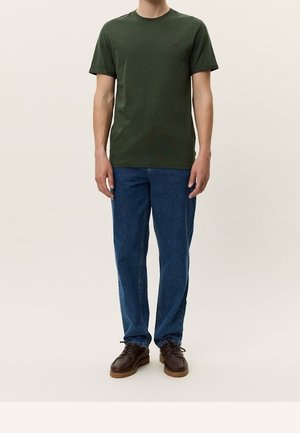Mężczyzna ubrany w ciemnozielony t-shirt z krótkim rękawem, niebieskie dżinsy i brązowe buty sznurowane, stojący na jednolitym tle.