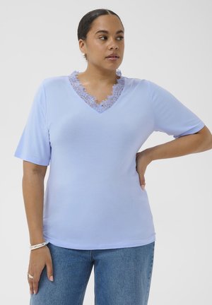 Vrouw draagt een lichtpaarse V-hals shirt met kanten rand en blauwe spijkerbroek, staand met één hand op haar heup en kijkt opzij.