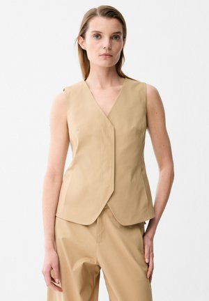 Donna che indossa un gilet beige senza maniche con scollo a V e pantaloni beige abbinati, in piedi davanti a uno sfondo bianco uniforme.