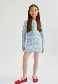 Set di cardigan e gonna a maglia azzurro chiaro con motivi testurizzati, abbinati a scarpe rosse basse e collant bianchi. Bottoni e tasche sull'outfit.