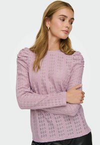 Kvinne med langt blondt hår iført en lavendel blonde teksturert langermet topp og små gullringøredobber, med armene i krysset, ser mot siden.