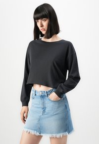 Woman with straight dark hair wearing a black long-sleeve cropped top and a light blue denim skirt with a frayed hem.