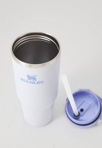 White Stanley metal tumbler with open top, stainless steel rim, and detached blue lid with white straw on white surface.