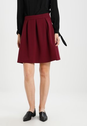 Kvinde iført et knælængde bordeaux plisseret nederdel, sort bluse med lange ærmer og sorte spidse flade sko, stående på en hvid baggrund.