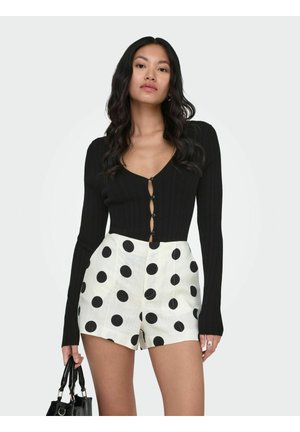 Femme portant un haut noir à manches longues boutonné et un short blanc taille haute à gros pois noirs, tenant un sac à main noir.