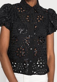 Blouse noire à manches courtes avec un motif en dentelle découpé floral, devant boutonné et manches bouffantes, portée avec un pantalon blanc.