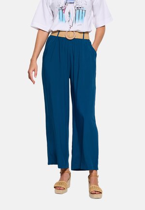 Mujer con pantalones anchos azules, cinturón trenzado color canela y sandalias de cuña estilo espadrille color canela, combinados con una camiseta gráfica blanca.