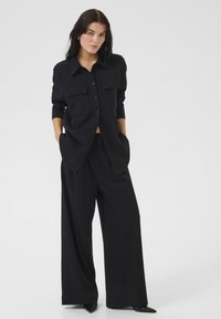 Zwarte oversized shirt met twee voorzakken, knoopsluiting en lange mouwen, gecombineerd met zwarte wijde broek. Gladde textuur.