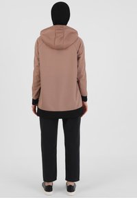 Chaqueta marrón con capucha, detalles negros, mangas largas y corte relajado. Combinada con pantalones negros y zapatos negros, con un fondo neutro.