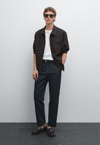 Giacca nera con colletto, t-shirt bianca, pantaloni scuri e mocassini neri. Gli accessori includono occhiali da sole scuri. Tessuto morbido, vestibilità su misura.