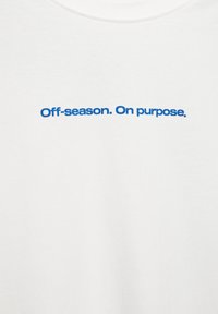 Biała bawełniana koszulka z niebieskim napisem „Poza sezonem. Celowo.” Materiał ma gładką fakturę i klasyczny dekolt typu crew neck.