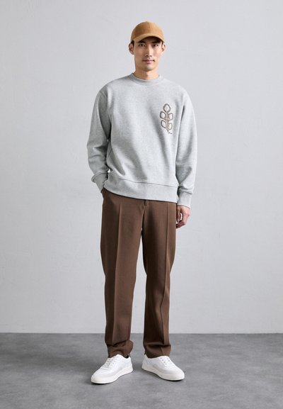 Felpa grigia con polsini a costine, design del logo marrone, abbinata a pantaloni marroni e sneakers bianche. Il modello è in piedi davanti a uno sfondo neutro.