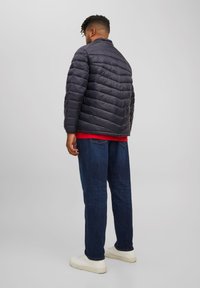 Chaqueta acolchada gris oscuro con acolchado horizontal, cuello alto y cierre de cremallera, llevada sobre una camiseta roja, combinada con jeans azules y zapatillas blancas.