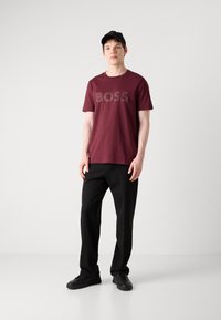 Burgundská bavlnená tričko s logom "BOSS", skombinované s čiernymi nohavicami a čiernymi topánkami. Model stojí na jednofarebnom bielom pozadí.