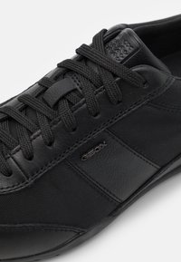 Black sneaker made of leather and textile, featuring a mesh panel, textured accents, and flat laces. Logo visible on the side.