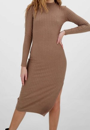 Woman wearing a long-sleeve, ribbed, knee-length brown dress with a side slit, standing with one hand on hip and the other relaxed.