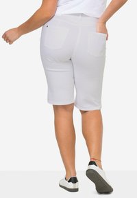 Shorts bermuda blancs au design ajusté, avec deux poches arrière, texture lisse et ourlet retroussé, associés à des baskets blanches et noires.