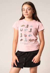 Lichtroze t-shirt met verschillende cartoonlaarzen, hoeden en een print van een sheriffster. Gecombineerd met een zwarte rok met knopen aan de voorkant.