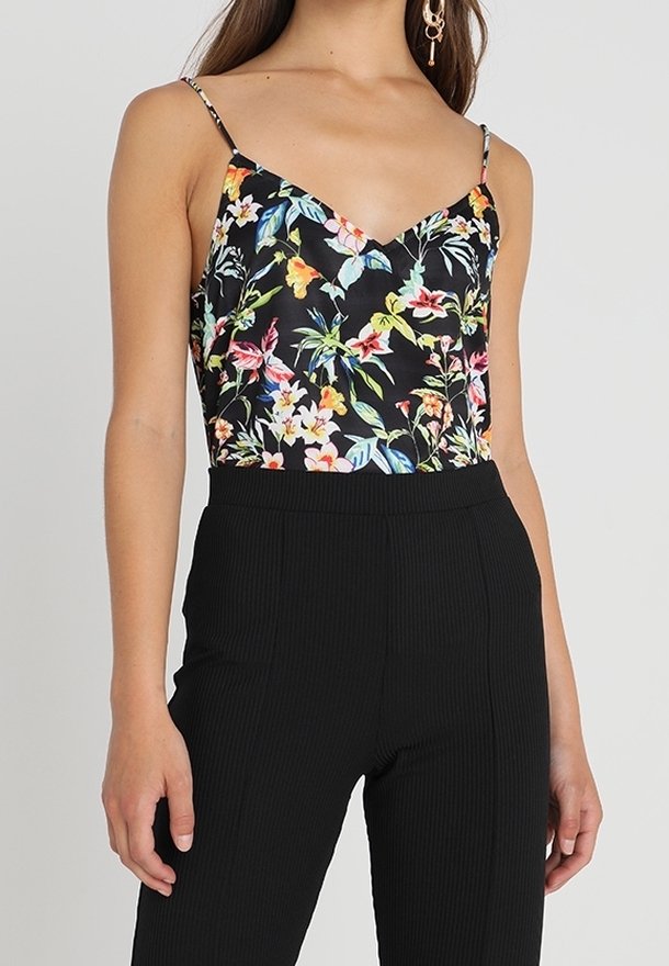 Femme vêtue d'un haut noir fleuri sans manches à col en V avec de fines bretelles, et d'un pantalon noir côtelé taille haute.