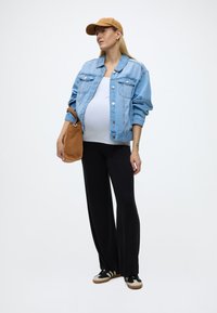 Raskaana oleva nainen, jolla on vaaleansininen farkkutakki, valkoinen paita, mustat housut, ruskea lippis ja joka pitää ruskeaa laukkua, seisoo yksiväristä taustaa vasten.