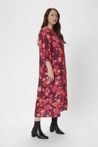 Robe longue arborant un motif floral dans des teintes de rouge et de rose, avec une coupe décontractée, des manches 3/4, et associée à des bottes à talons noires.