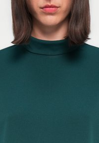 Top teal a collo high realizzato in tessuto liscio, caratterizzato da un design aderente e da una delicata cucitura decorativa. La texture sembra morbida e lucida.
