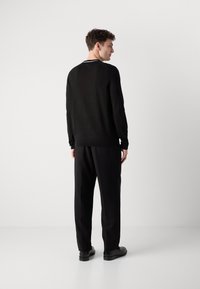 Svart strikket langermet skjorte med kontrasterende hvit stripe ved kragen, kombinert med løse svarte bukser og svarte sko.