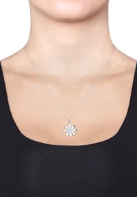 Sølv halskæde med en floral vedhæng, der har lagdelte kronblade og en central sten, vist på en sort top mod en neutral baggrund.