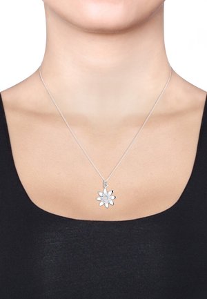 Silberkette mit einem floralen Anhänger, der geschichtete Blütenblätter und einen zentralen Stein zeigt, auf einem schwarzen Oberteil vor einem neutralen Hintergrund präsentiert.