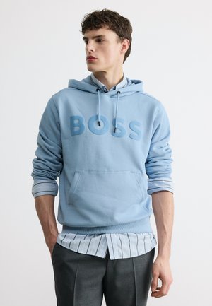 Noor mees, kes kannab helesinist kapuutsiga pusa, selle all triibulist nööbitavat särki ja tumedaid halli pükse, vaadates vasakule.