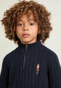 Tumesinine tõmblukuülikond, millel on ribatekstuur ja kootud nöördisain. Rinnal on väike broderi nutcracker detail.