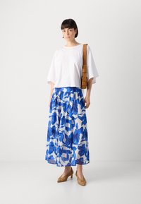 Haut court blanc avec des manches larges, associé à une longue jupe florale bleue. Un sac à main en cuir marron et des chaussures à talons tressées complètent le look.