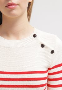 Pull blanc en maille avec des rayures rouges horizontales. Il comporte trois boutons noirs sur l'épaule. Texture lisse et col rond.
