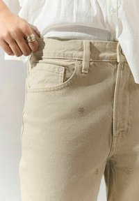 Jeans en coton beige présentés à la taille, avec des poches avant, une passante de ceinture et des motifs floraux brodés décoratifs.