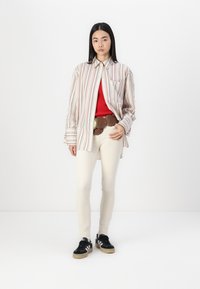 Striped long-sleeve shirt in cream, red, and navy, layered over a red top. Slim white pants and black sneakers with beige soles. Brown belt with metal accents.