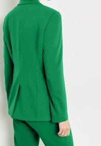 Blazer verde a medida con hombros estructurados, una abertura en la parte trasera y textura de tela suave, llevado sobre pantalones a juego.