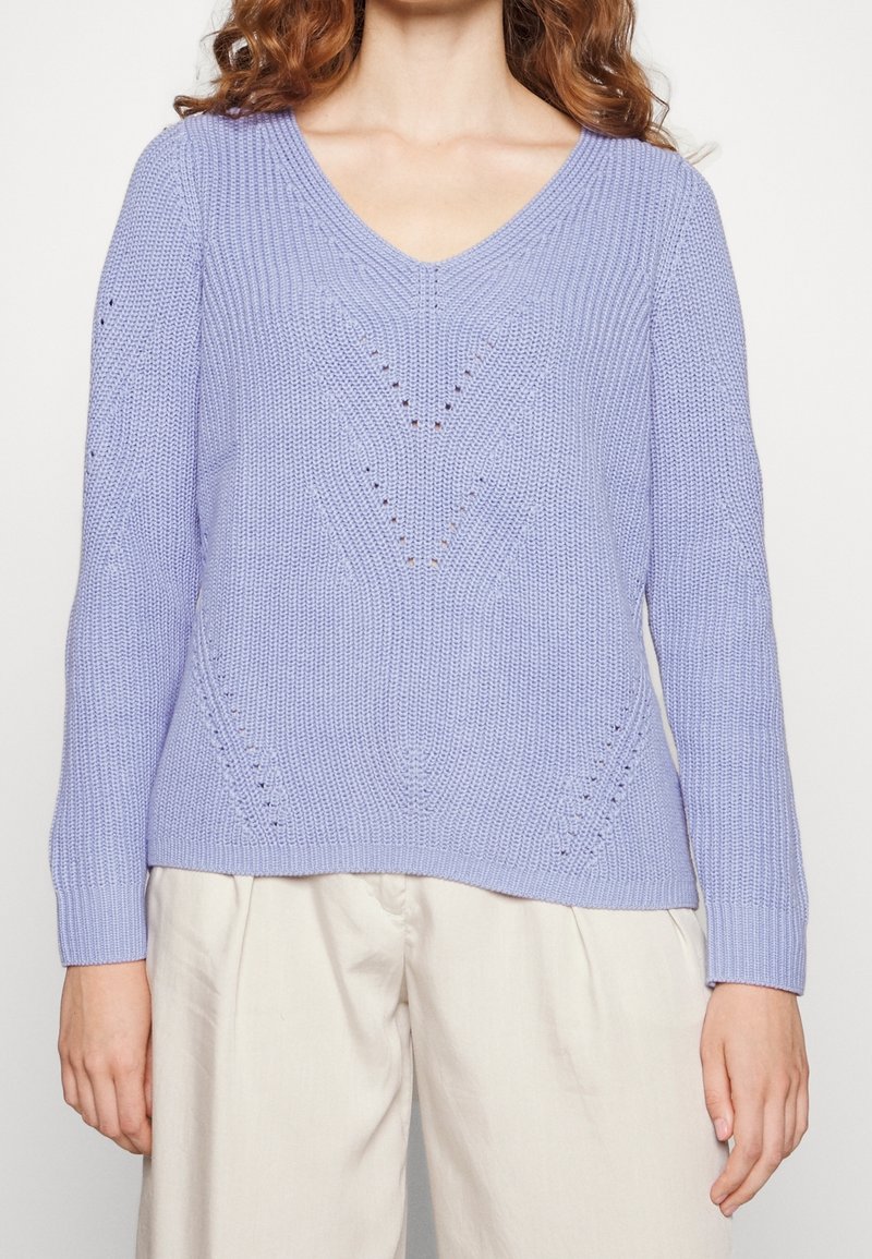 Femme portant un pull en tricot violet clair à col en V avec des motifs en chevrons discrets, associé à un pantalon ample de couleur crème.