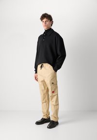 Svart oversized polo-skjorte med knapper, kombinert med beige cargo-bukser med sidelommer og en rød merkelapp, fullført med svarte sko.