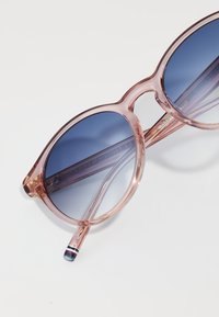 Lunettes de soleil rondes avec monture transparentes roses et verres dégradés bleus. Présente des accents dorés subtils et des embouts de branches rayés.