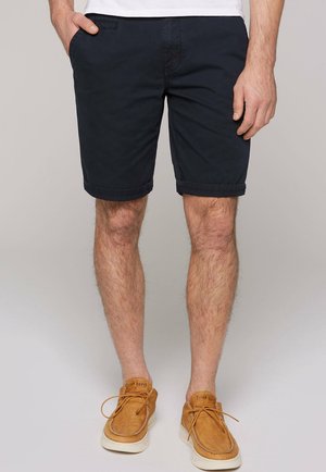 Pantalones cortos de algodón azul marino con un diseño clásico, dobladillos enrollados y bolsillos laterales. Combinados con zapatos casuales color beige y una camiseta blanca lisa.