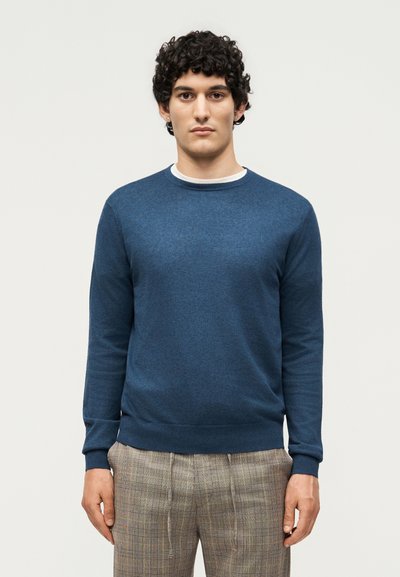 Hombre con cabello rizado, vestido con un suéter azul de cuello redondo y pantalones de cuadros, de pie contra un fondo claro y liso.