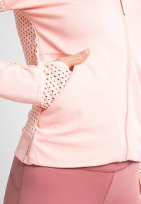 Hand resting in pocket of light pink jacket with perforated cuff details, paired with mauve leggings.