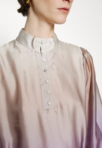 Blouse en soie avec un col montant, une patte de boutonnage et un doux dégradé du beige clair au violet ; des manches bouffantes ajoutent une texture subtile.