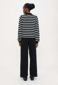 Kvinne med fletteknute, iført svart-hvitt stripete genser, vide svarte bukser og svarte støvler, stående med ryggen mot på hvit bakgrunn.