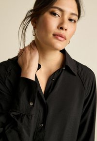 Chemise noire à col fabriquée en tissu léger avec des boutons brillants, présentant une coupe décontractée et des détails de couture visibles sur les poignets.