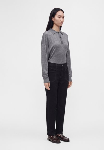 Femme aux longs cheveux noirs portant un polo gris à manches longues, un pantalon noir et des mocassins marron foncé, debout devant un fond blanc uni.