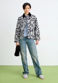 Persona che indossa un giubbotto floreale nero e grigio, una camicia blu, jeans larghi e sneakers lilla, mentre tiene una borsa nera pelosa su un pavimento verde.